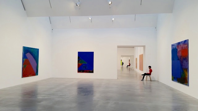 John Bingham working at the John Hoyland Show - John seated as an invigilator in the studio space with three large pieces by John Hoyland, all abstract art in blues, reds and violet colours.