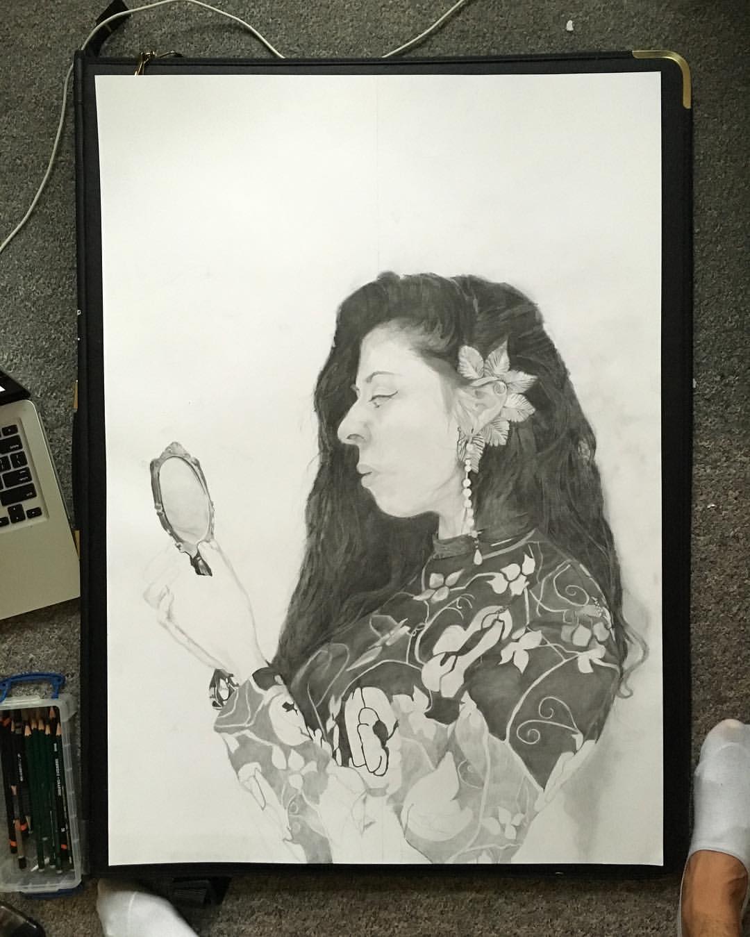 John Bingham's portrait in progress in black and white pencil of a woman with a floral jumper on, long wavy hair, a flower behind her ear and looking into a small elaborate mirror