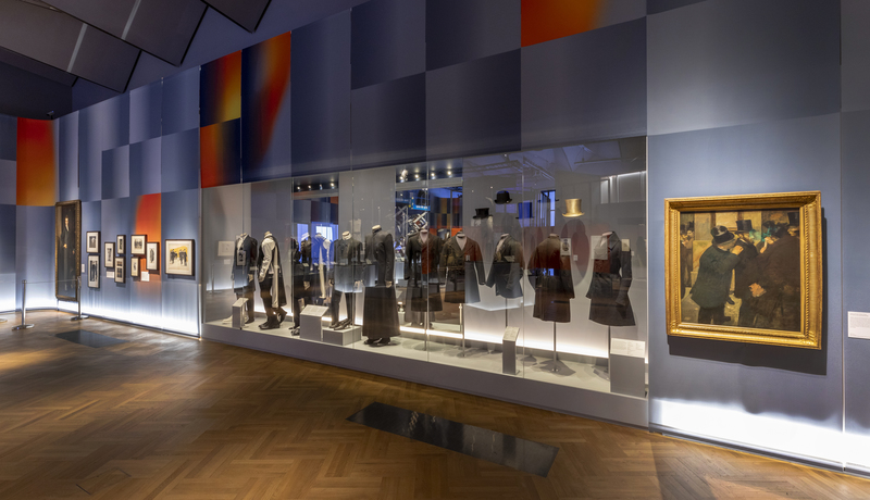 Gallery of mannequins dressed in menswear on plinth behind glass with painting on right hand side depicting historic scene of males gathering in suits