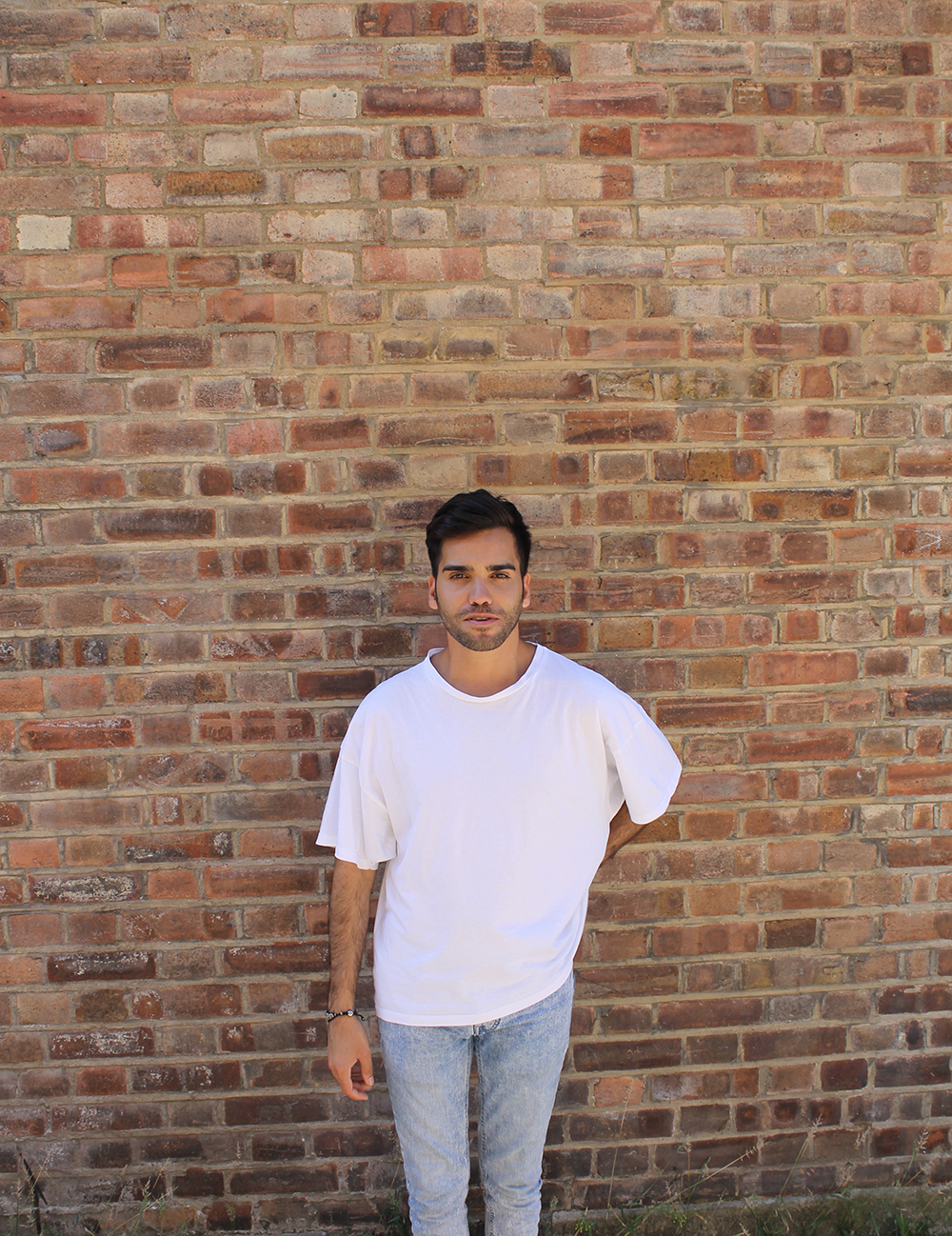 John Bingham in a white t-shirt and faded jeans standing in front of a red brick wall