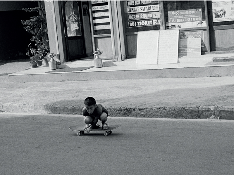 Photograph by Tom Caron-Delion, featured in a joint exhibition Thik Cha: An Exhibition for Nepal. The black and white photograph is of a young Nepalese boy crouching down on his skateboard on an empty road in front of a shop with many signs and potted plants.