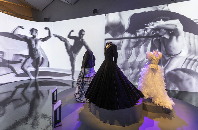 3 mannequins in ballgowns, 2 have male jackets. Galley backdrop is of male dancers in muscular poses