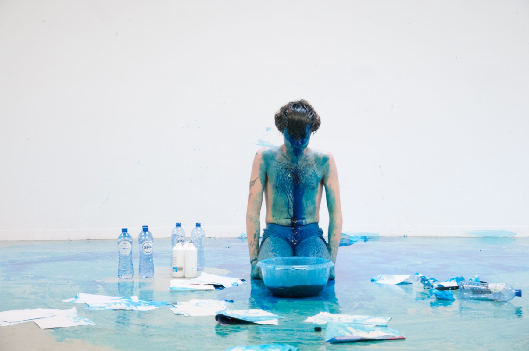 Giovanni kneels in a white room with a bowl of blue ink in front of him, he wears no shirt and is covered in blue ink diluted by water surrounded by paper, bottles of water and the blue ink.