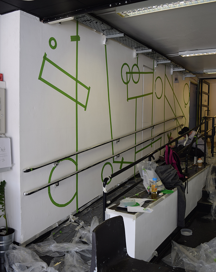 The left hand side wall in white with green masking tape placed for the geometric shapes
