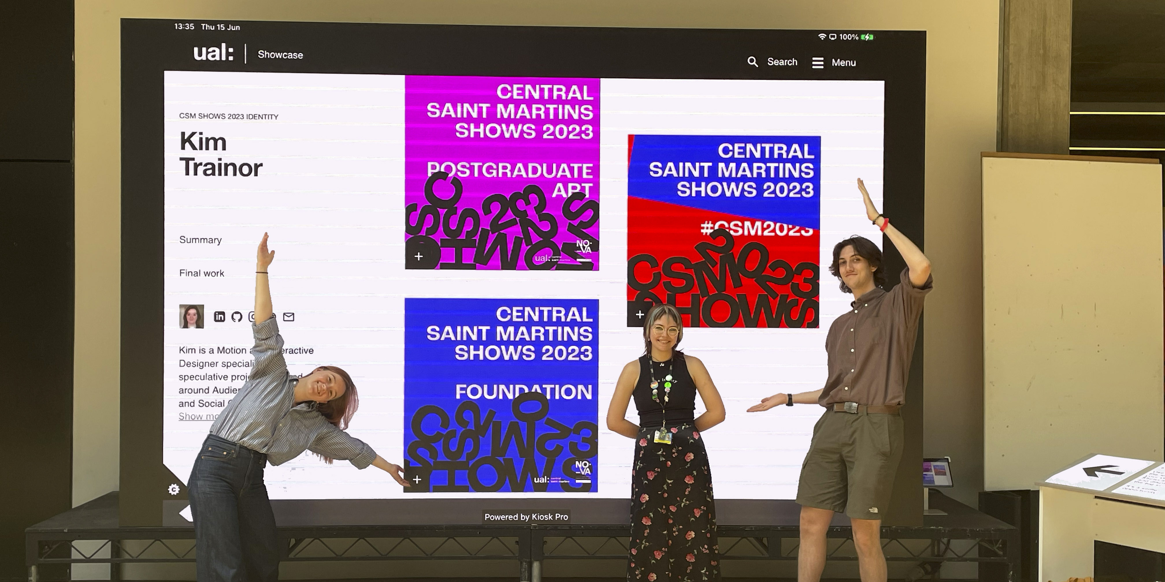 Three students stand happily in front of a large display screen