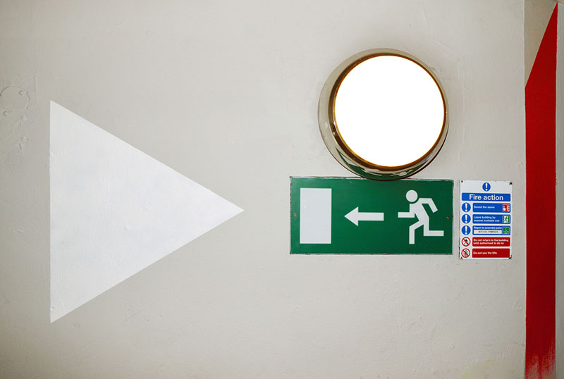 Debruised by Kimathi Donkor 2015 mural installation, 1m x 10 m white triangular sign next to an exit sign on a white wall