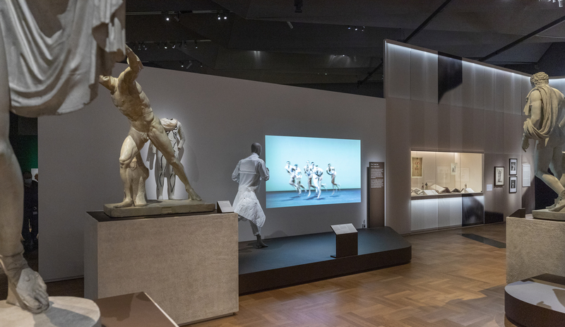 male mannequin running against a screen backdrop in a gallery surrounded by naked male statues