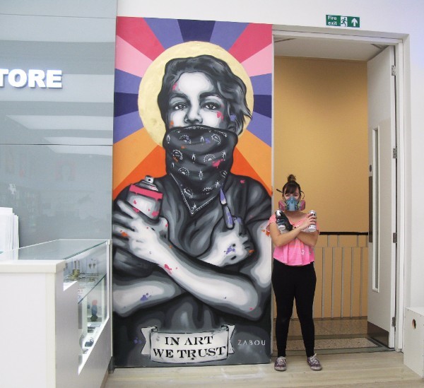 'IN ART WE TRUST' is a spray painted piece by street art talent Zabou who stands next to her work in this image. The piece in Saatchi Store is of a young person with a bandanna covering their mouth, at the bottom is a banner saying "IN ART WE TRUST". The young person's arms are crossed over their chest, in one hand is a spray paint can, in the other is a paintbrush. The image is mostly grey, black and white except for blobs of paint scattered across the image. The young person has a yellow circle behind their head which looks like a halo, lines of orange, purple, blue and pink radiate out from this yellow circle.