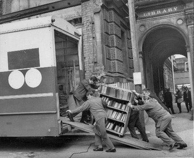Black and white photograph of removal men putting a trolley of books into a van