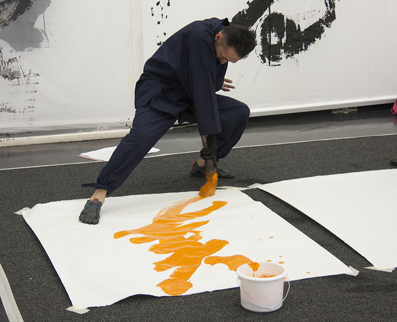 Japanese artist Houran Yokoyama paints a calligraphy word using a big brush, yellow paint on a big white piece of paper placed on the floor